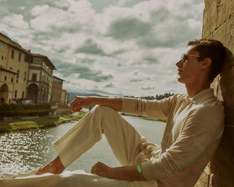 A young man wearing sunglasses and casual wear sits on a stone ledge along a river in an old European city under a partly cloudy sky.