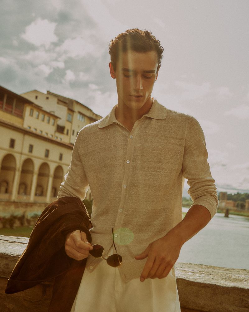 A stylish man in a polo shirt stands on a bridge in Florence, Italy, holding sunglasses, with buildings and a river in the background.