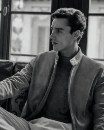 Carlos Galobart seated indoors wearing a suede zip jacket and crew-neck top for Brett Johnson.