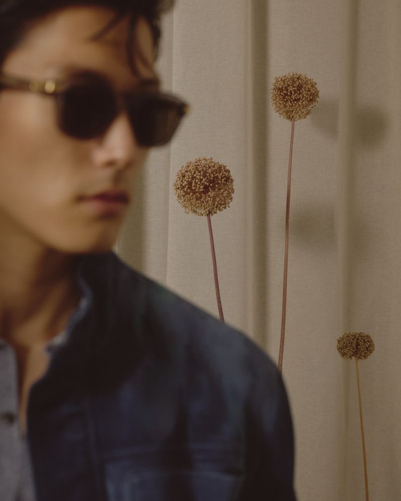 Portrait of an Asian adult male with sunglasses, wearing a denim jacket, standing near dried flower stems against a neutral backdrop.