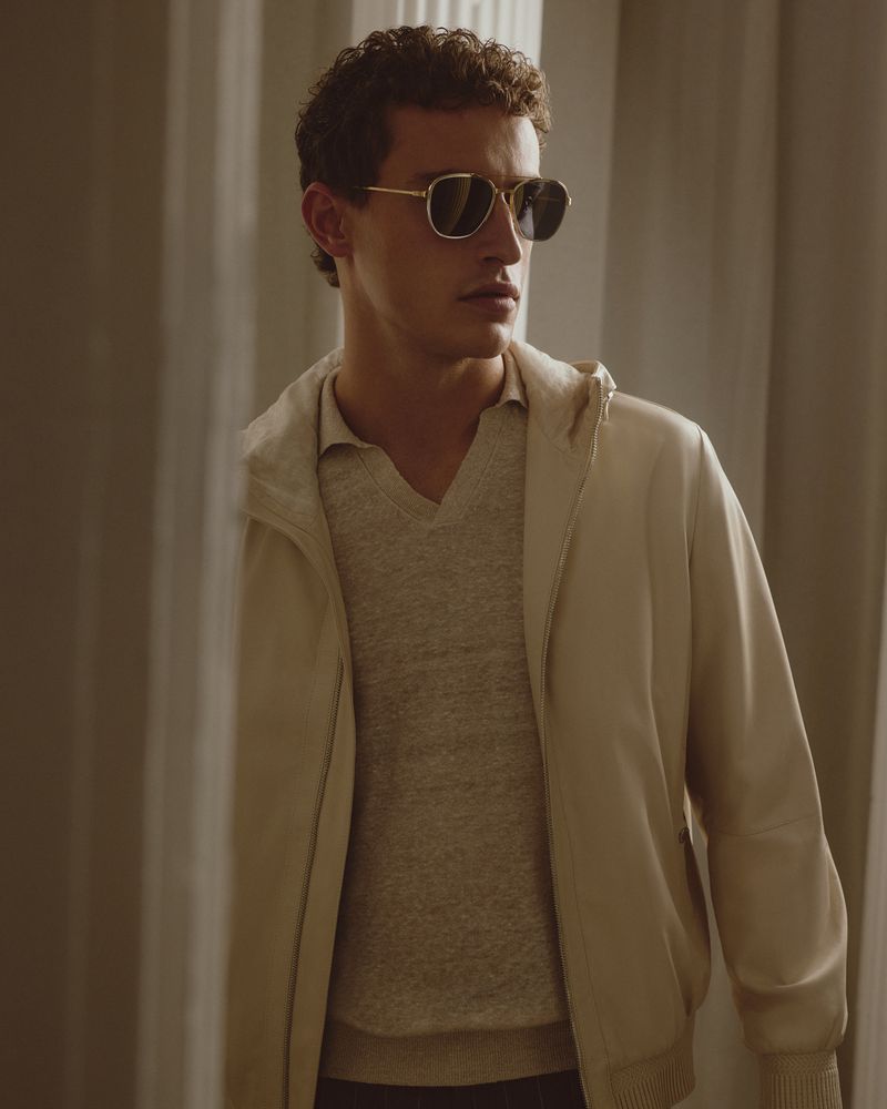 An adult male poses indoors wearing sunglasses, a light-colored jacket, and a sweater, standing in front of neutral-toned curtains.