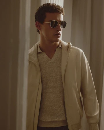 An adult male poses indoors wearing sunglasses, a light-colored jacket, and a sweater, standing in front of neutral-toned curtains.