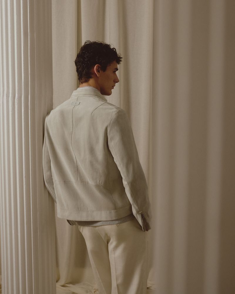 Back view of a young adult man posing in a light jacket and pants near a column and curtain, looking away, indoor studio shot.