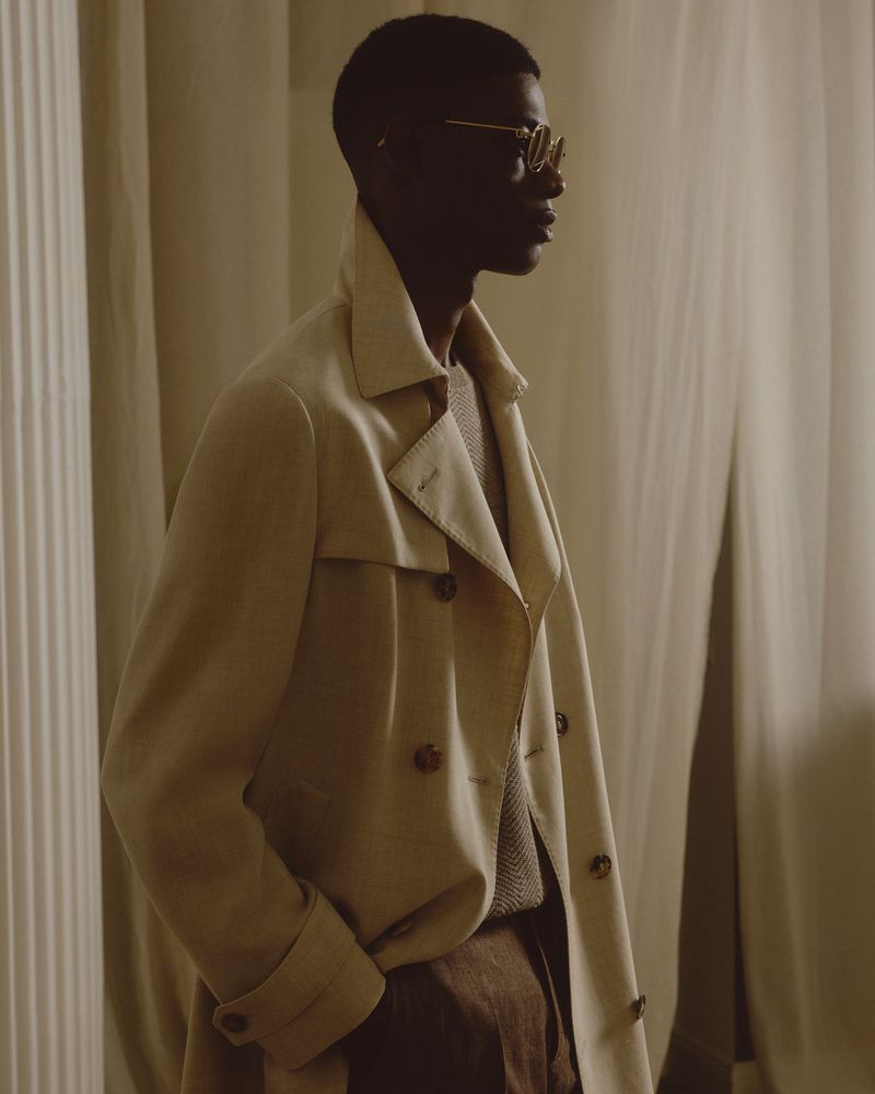 An adult male wearing a beige trench coat and glasses stands indoors near a column and curtain, photographed in a medium shot.