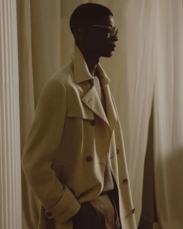 An adult male wearing a beige trench coat and glasses stands indoors near a column and curtain, photographed in a medium shot.