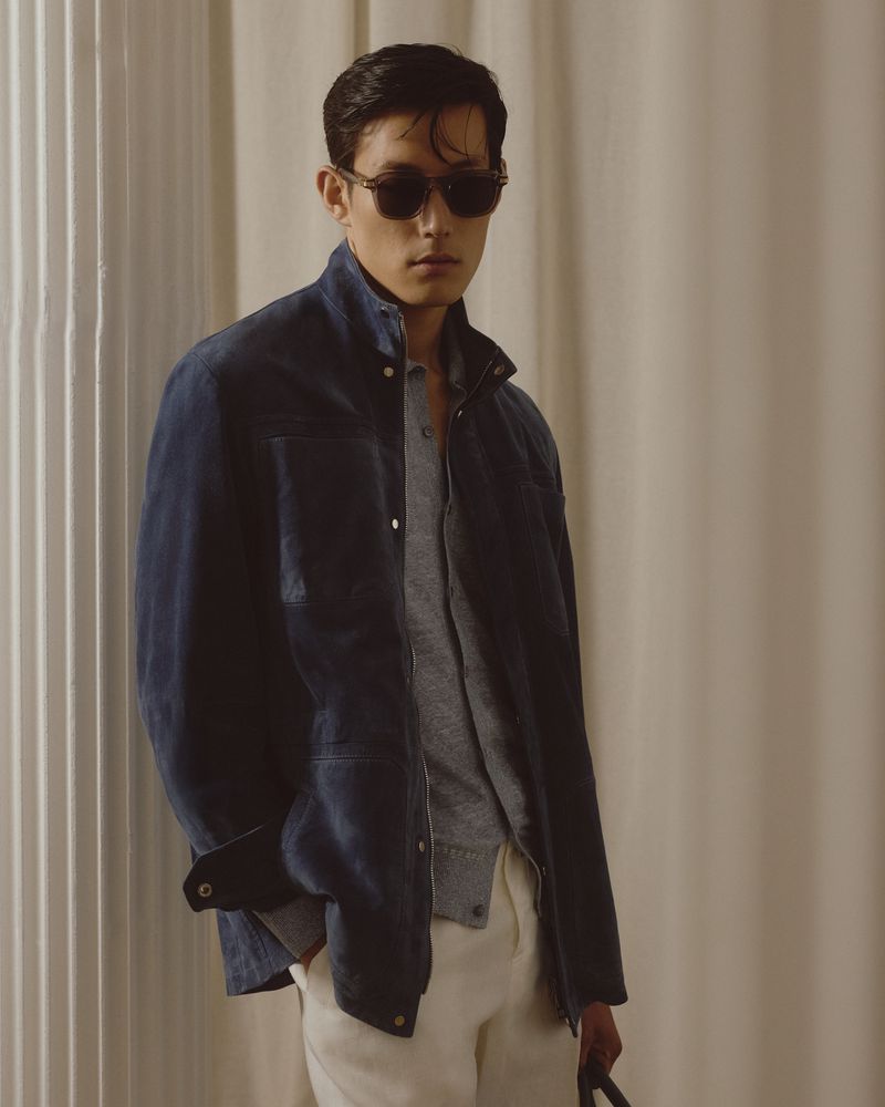 Portrait of an adult male wearing sunglasses, a blue jacket, and light pants, posing in an indoor environment with soft lighting.