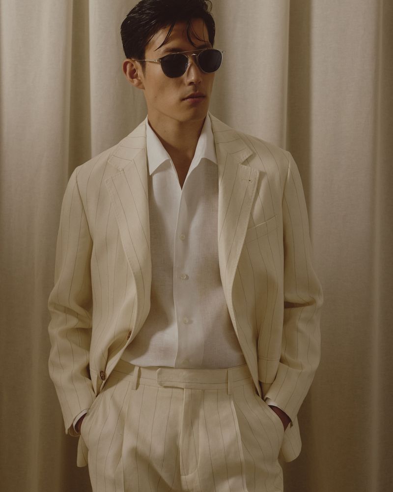 Man in light-colored striped suit and sunglasses poses against a tan fabric backdrop in a studio setting, looking away from camera.