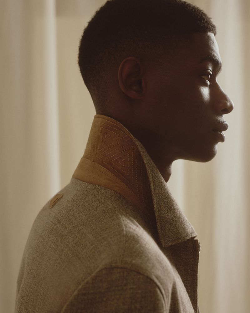 Profile portrait of an adult Black man with short hair, wearing a grey coat with tan collar, in front of a neutral-toned background.
