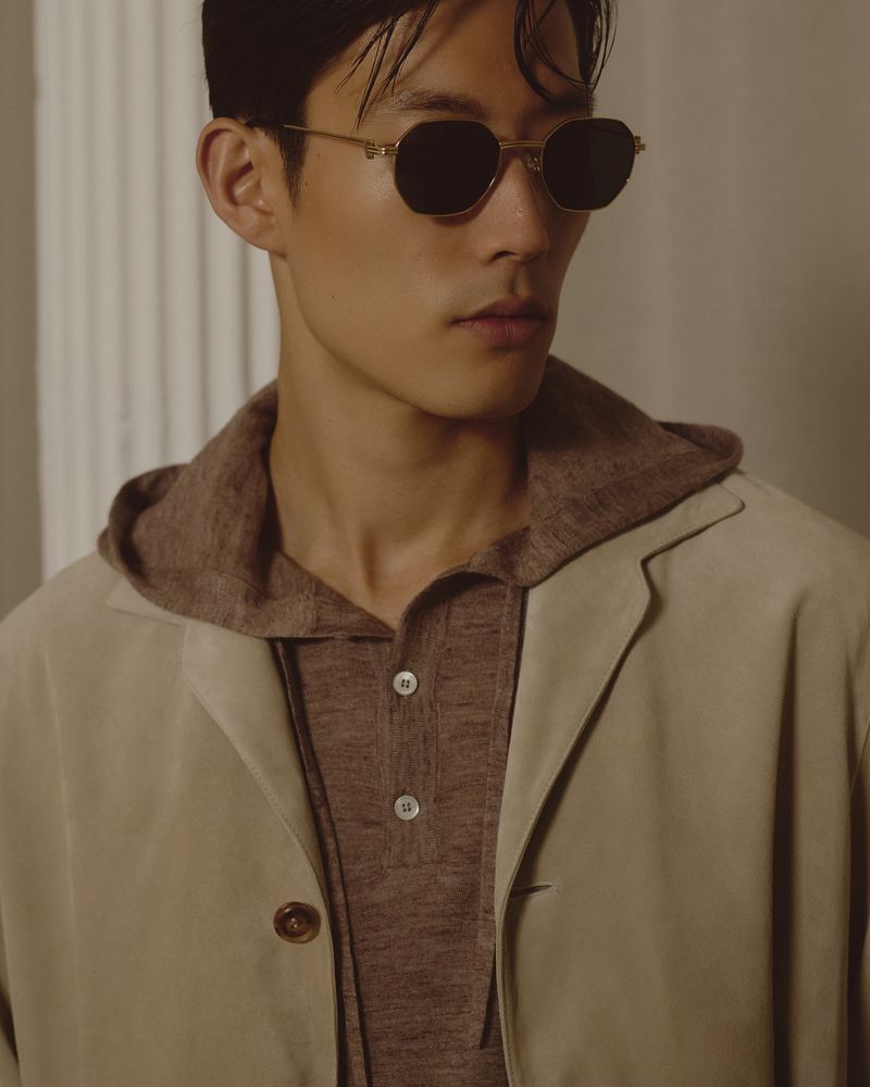 Asian man wearing sunglasses, a brown hoodie, and a beige jacket poses indoors against a neutral background in a medium shot.