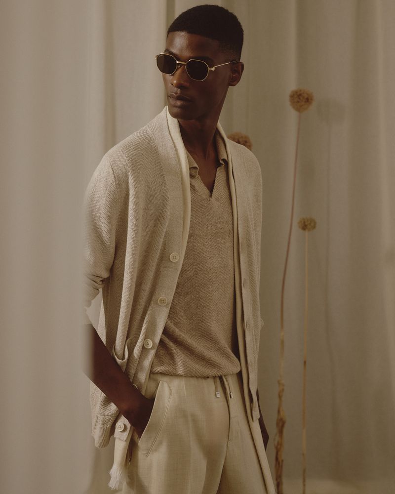 Portrait of a dark-skinned adult male wearing a beige knit cardigan, polo shirt, and trousers. He is also wearing sunglasses.