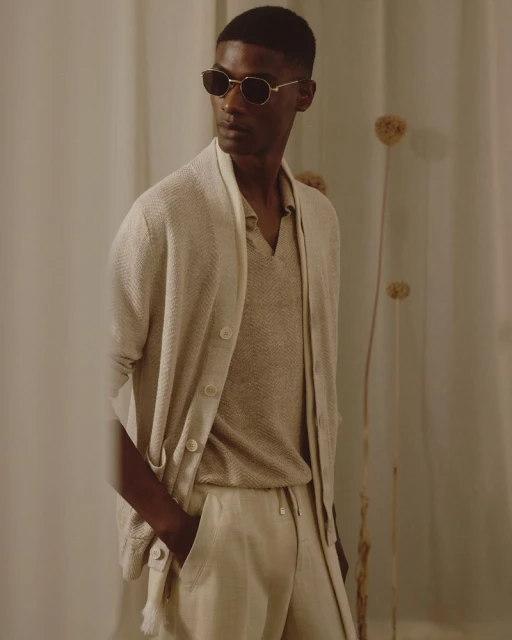 Portrait of a dark-skinned adult male wearing a beige knit cardigan, polo shirt, and trousers. He is also wearing sunglasses.