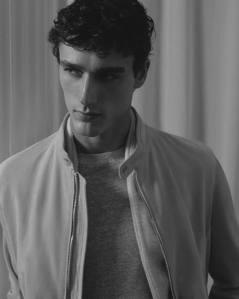 Monochrome portrait of a male fashion model with dark curly hair, wearing a light colored jacket and grey sweater in a studio.