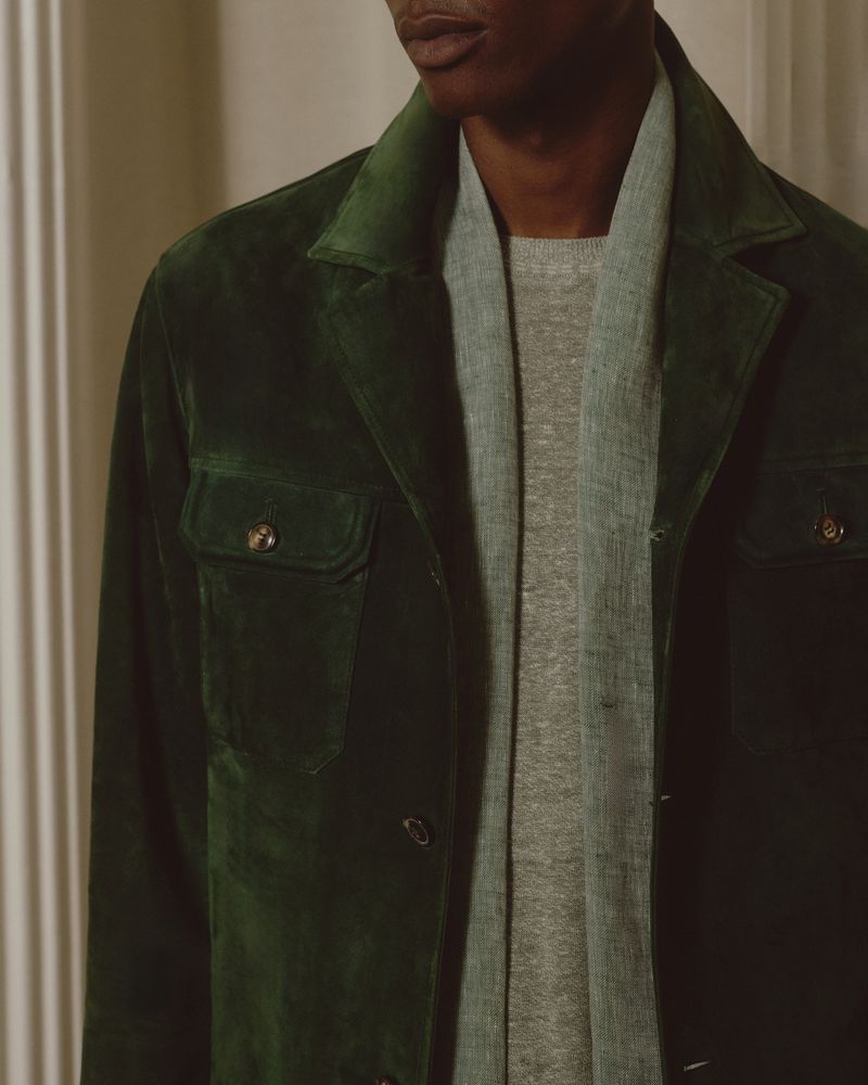 A stylish adult male poses indoors wearing a green suede jacket over a gray sweater and scarf. Neutral background with natural light.