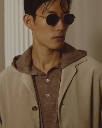 Model Juhyung Kang wearing dark geometric sunglasses, a brown knit hooded polo, and a beige suede jacket for Brett Johnson.