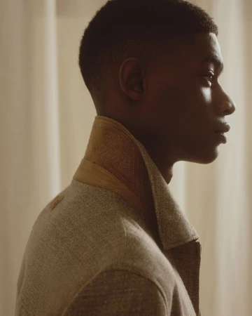 Profile view of a Black male model wearing a textured jacket with an upturned stitched collar against soft curtains.