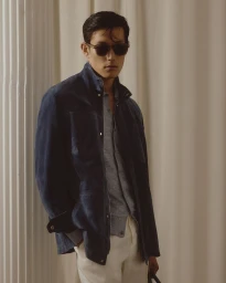 Model Juhyung Kang wearing dark sunglasses, a navy suede jacket, grey knit shirt, and cream trousers in an editorial setting.