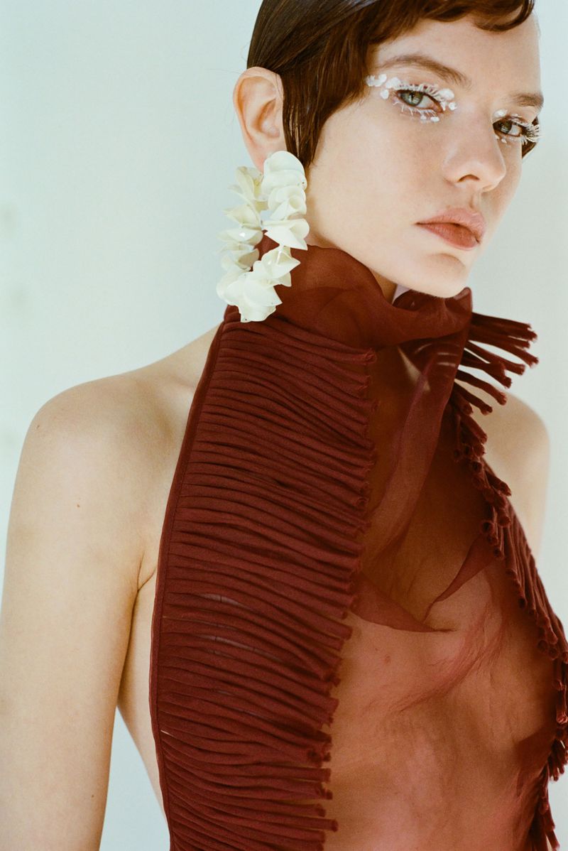 Woman with short hair, wearing large earrings and a reddish-brown dress, poses against a white background in a fashion studio setting.