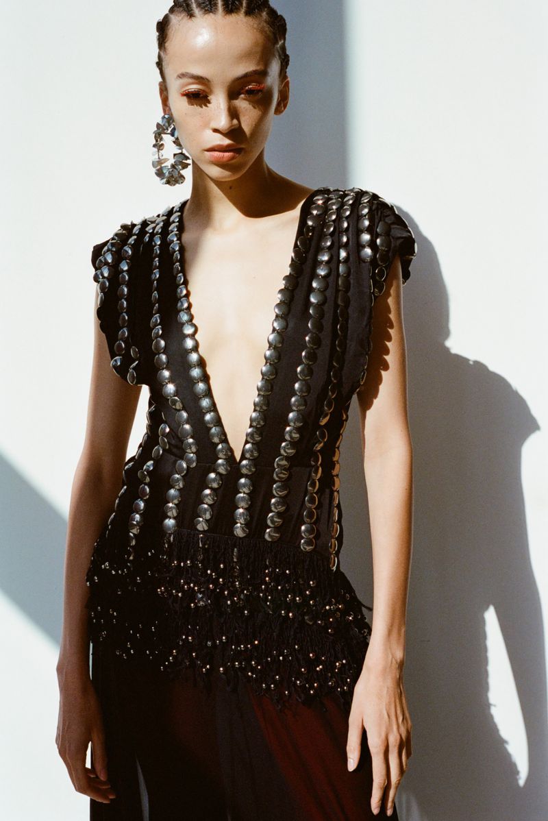 Fashion portrait of an adult woman with braids wearing a deep V-neck top with circular metallic embellishments and large disc earrings.