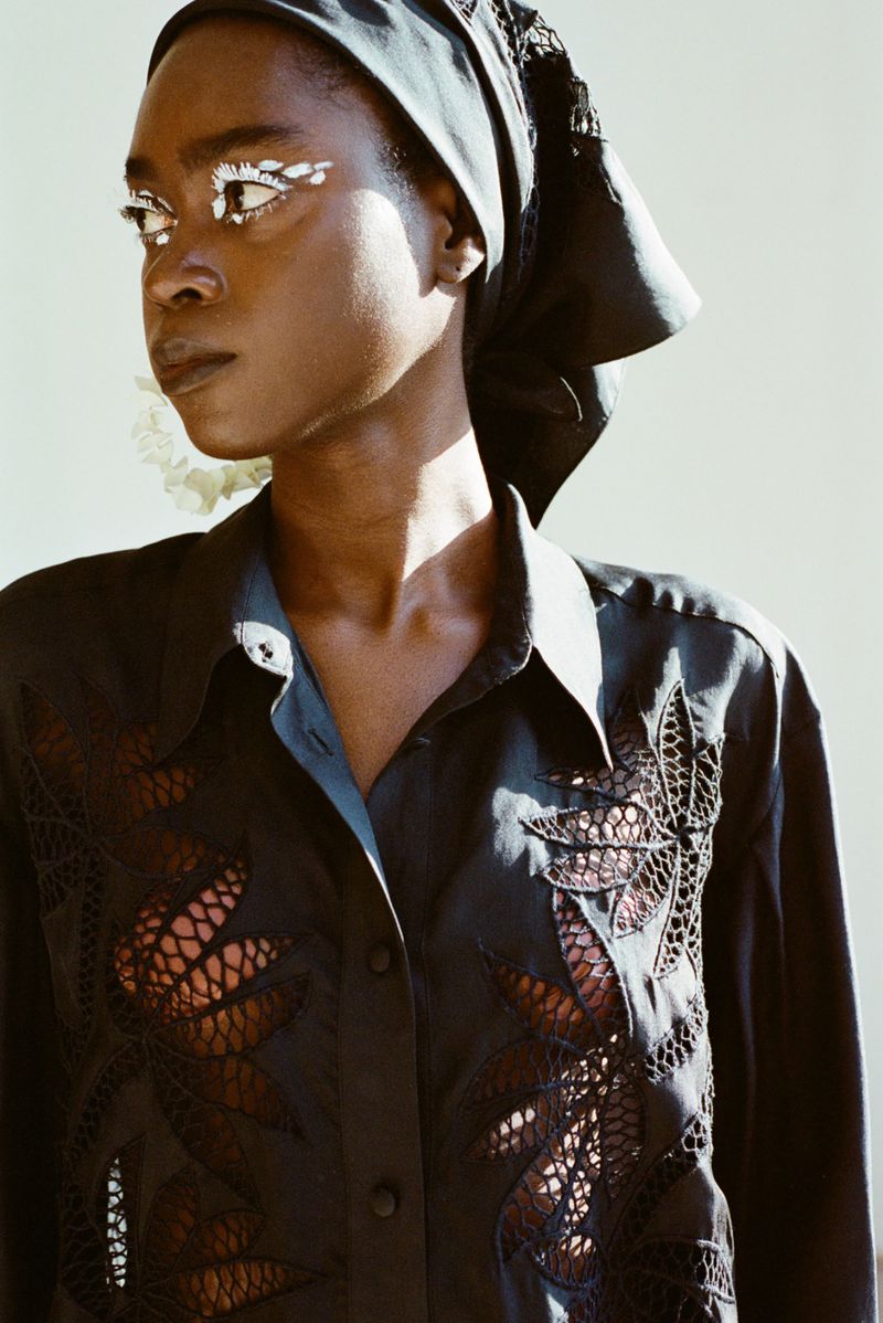 Woman with dark skin and bright makeup wears a headscarf and an openwork shirt in a studio shot with natural lighting.