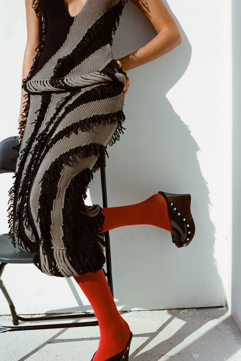 Fashionable adult female poses in a knitted dress with fringe detail, red stockings and black wedge heels against a white wall.