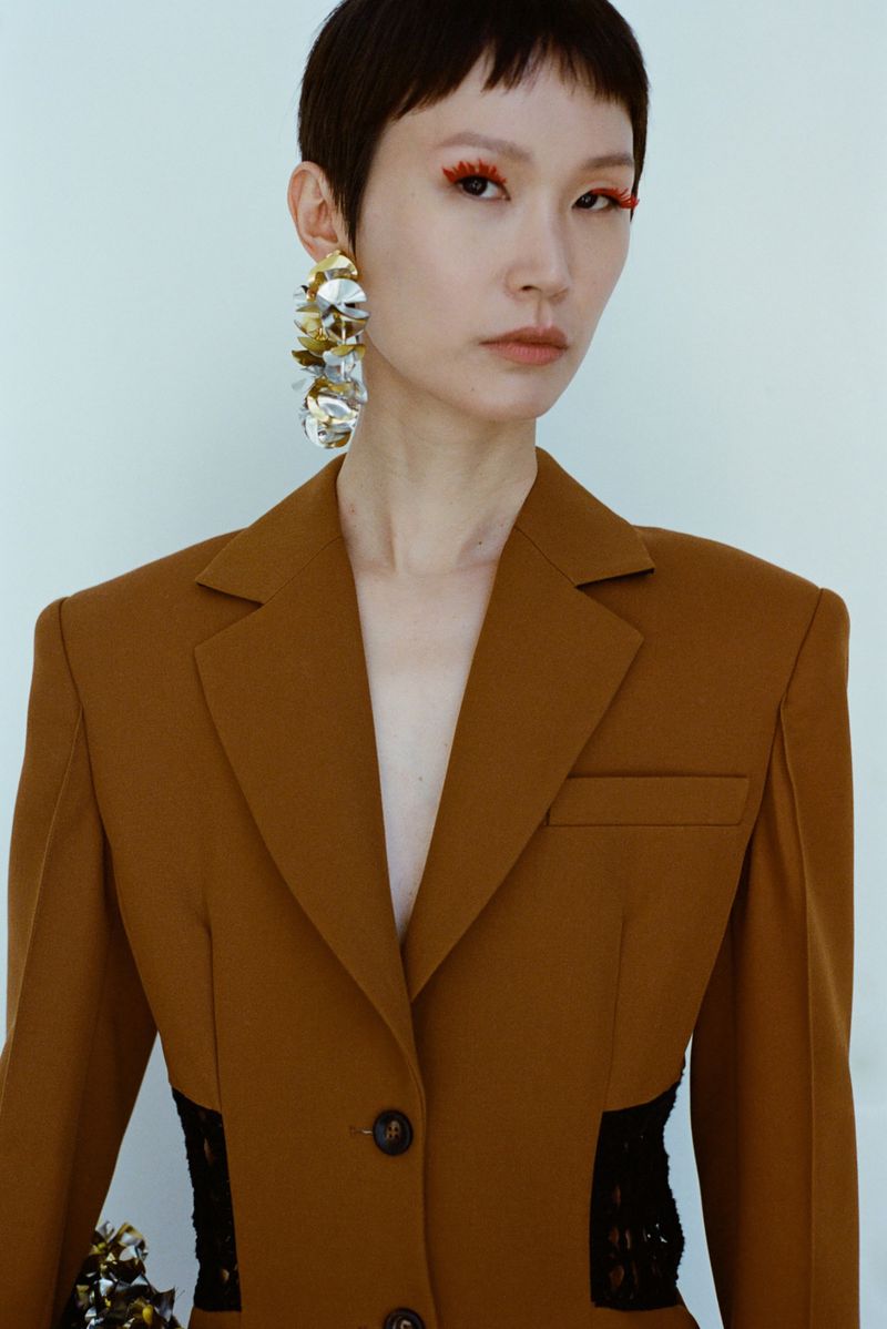 Close-up shot of a stylish Asian woman in a blazer with statement earrings and orange eyeliner, posing indoors against a white background.