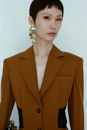 Close-up shot of a stylish Asian woman in a blazer with statement earrings and orange eyeliner, posing indoors against a white background.