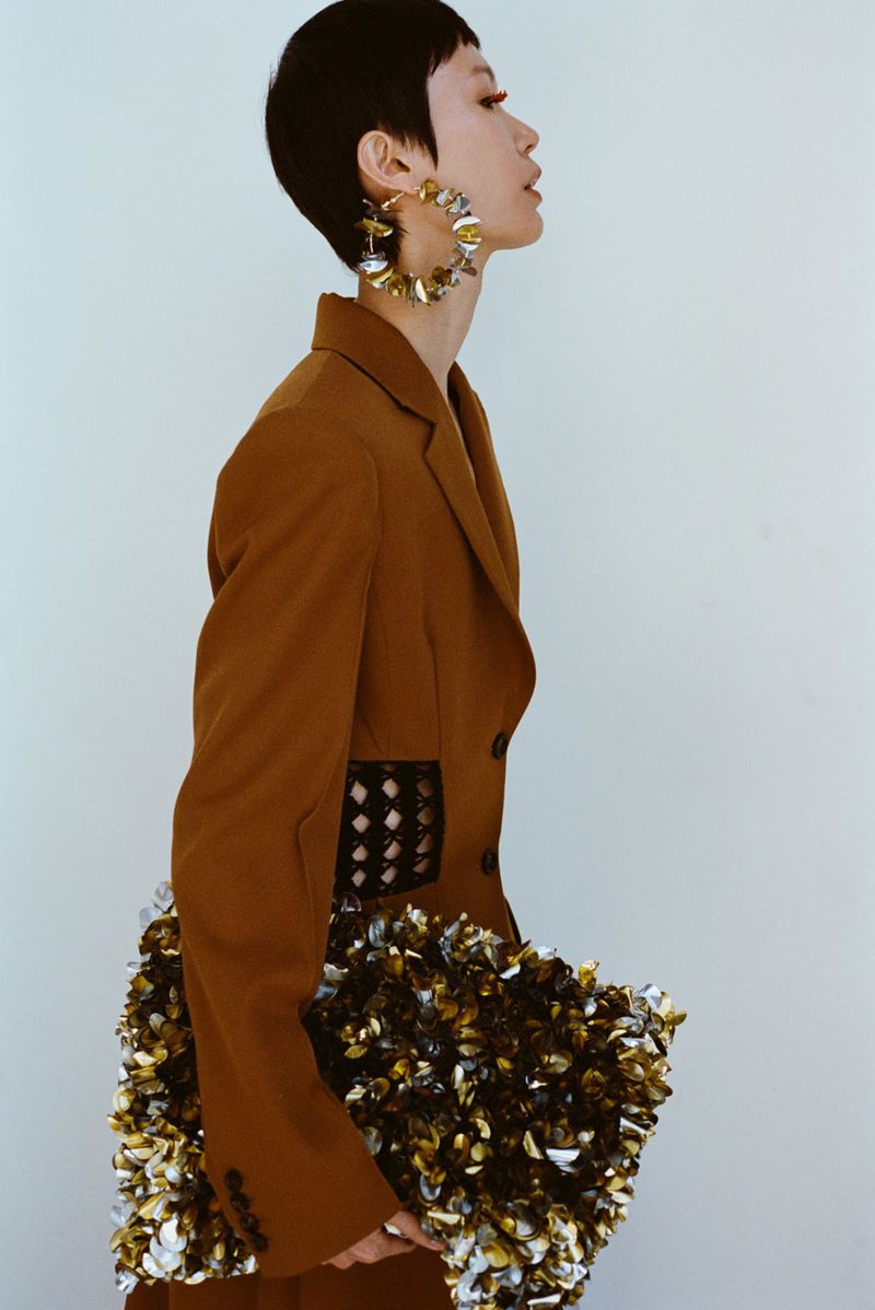 Profile portrait of an Asian model wearing a brown blazer, large earrings, and holding a shiny clutch in a studio setting.