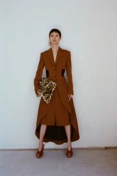 Model standing against a plain white wall wearing a long brown coat and holding a heavily textured gold handbag