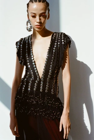 Model wearing a black embellished V-neck top with silver studs and braided hair against a white wall.