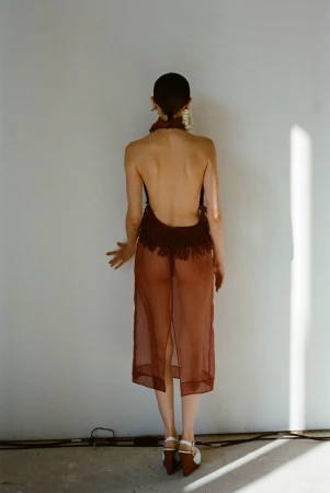 Model facing a white wall wearing a sheer brown halter dress with fringe detail and platform wedge shoes
