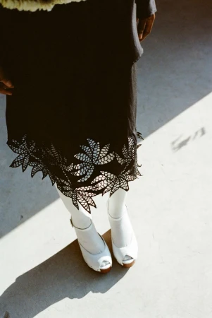 High-angle shot of a model wearing a black laser-cut skirt, white tights, and white platform peep-toe shoes.