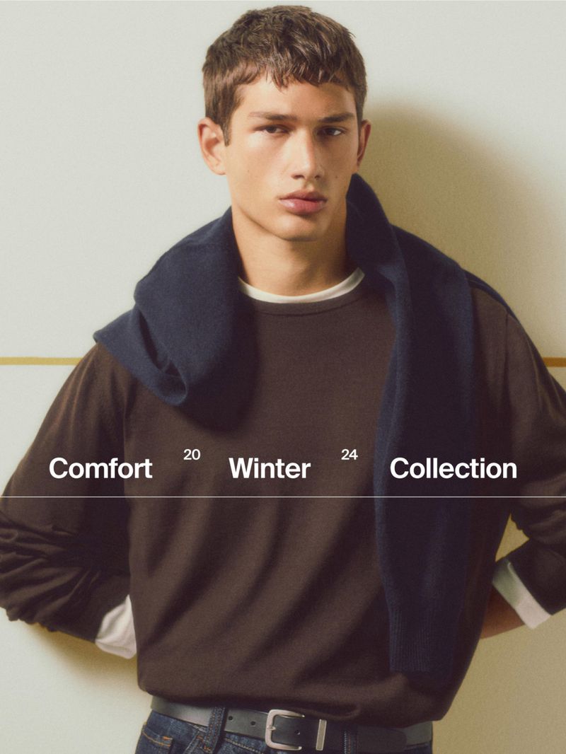 A young male model wearing a brown crewneck sweater with a dark blue sweater draped over his shoulders against a light wall