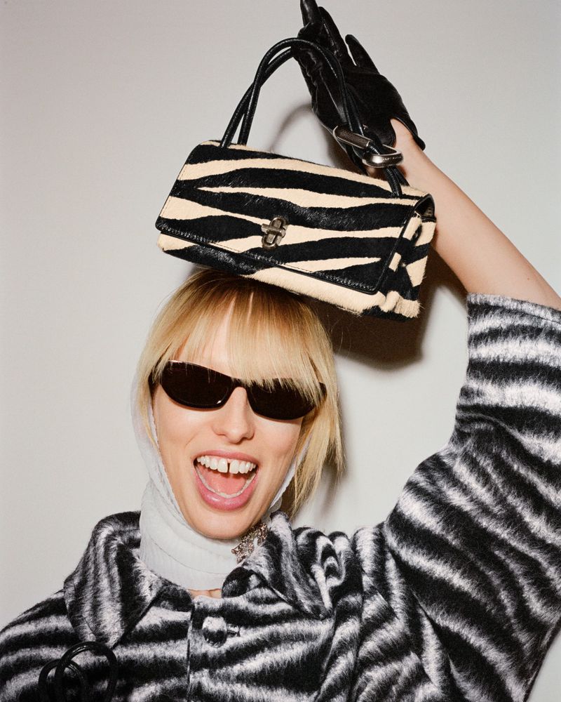 A blonde woman with sunglasses and a zebra-print coat holds a matching zebra-print handbag above her head indoors.