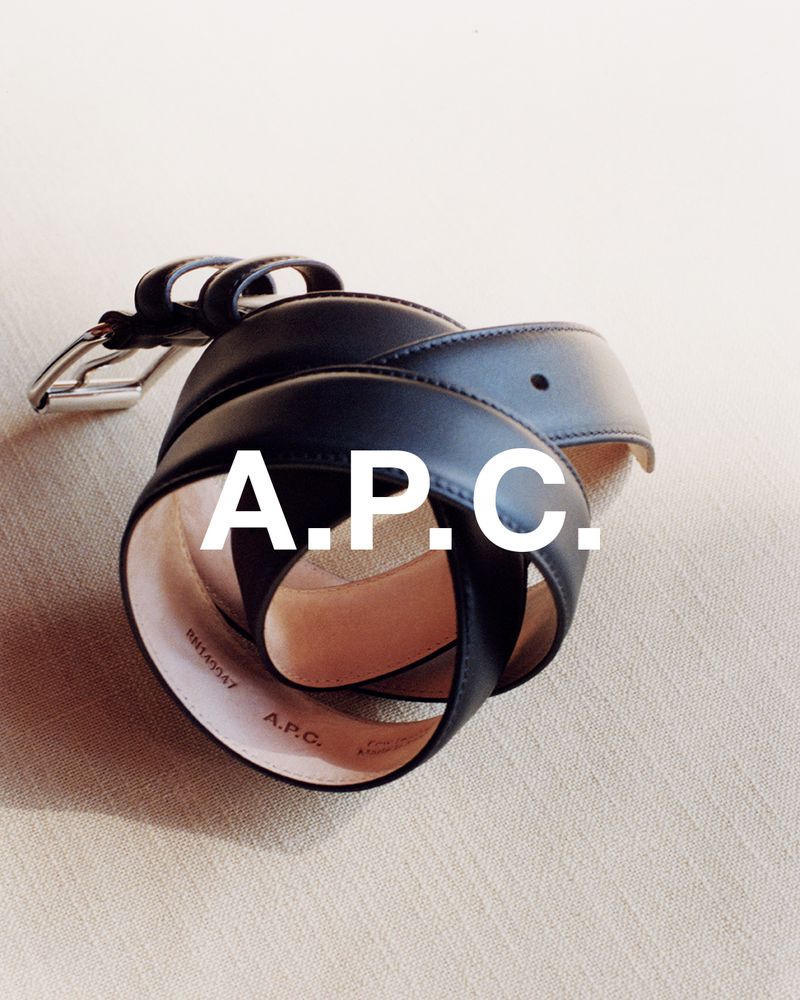 Overhead shot of black leather belt with silver buckle curled on textured beige surface with fashion logo APC in white text overlay.