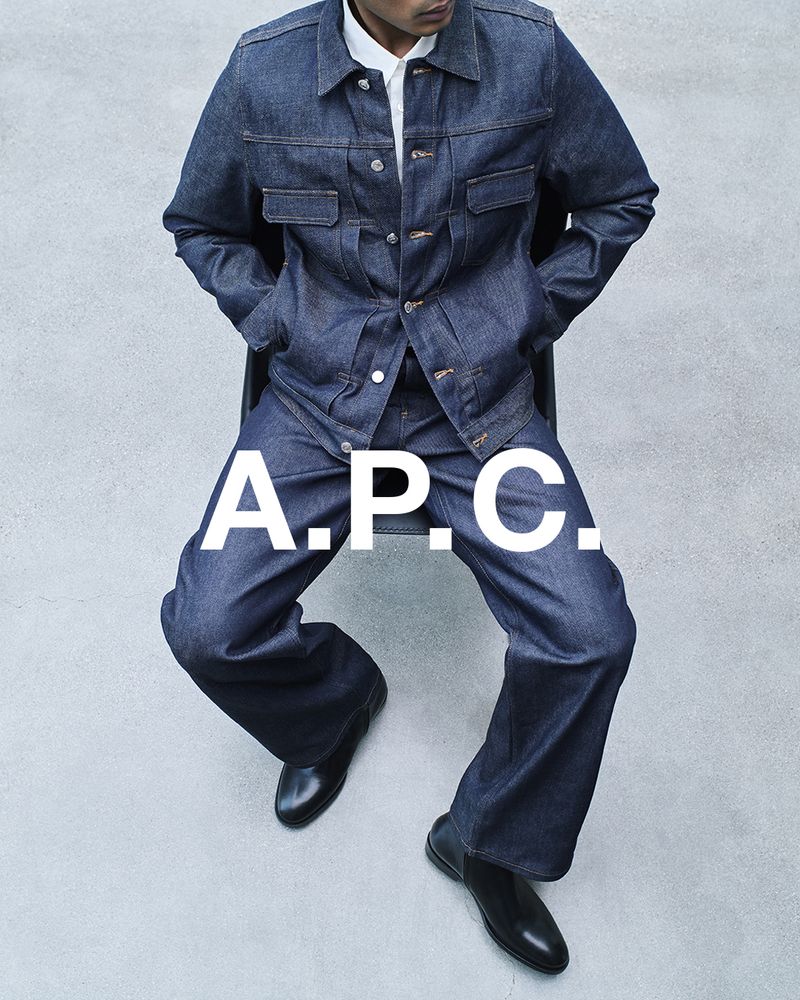 Man in dark denim jacket and pants sitting against grey backdrop, wearing black leather shoes. Large A.P.C. logo overlay.