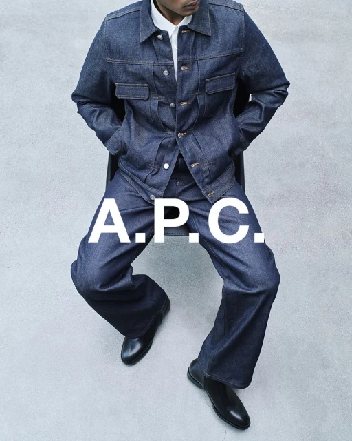 Man in dark denim jacket and pants sitting against grey backdrop, wearing black leather shoes. Large A.P.C. logo overlay.