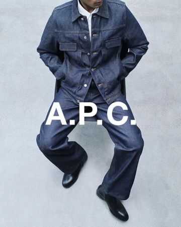 Man in dark denim jacket and pants sitting against grey backdrop, wearing black leather shoes. Large A.P.C. logo overlay.