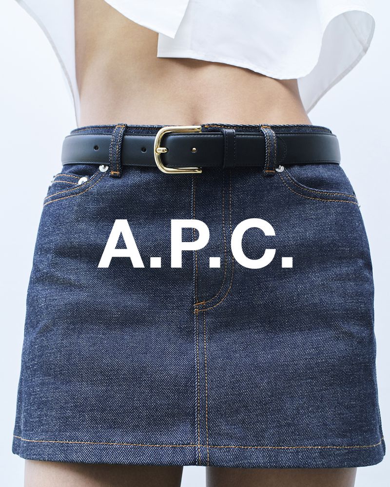 Close-up of a woman wearing a denim miniskirt with a black belt and a white top against a white background. Brand logo on the skirt.