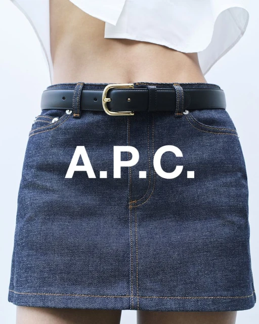 Close-up of a woman wearing a denim miniskirt with a black belt and a white top against a white background. Brand logo on the skirt.