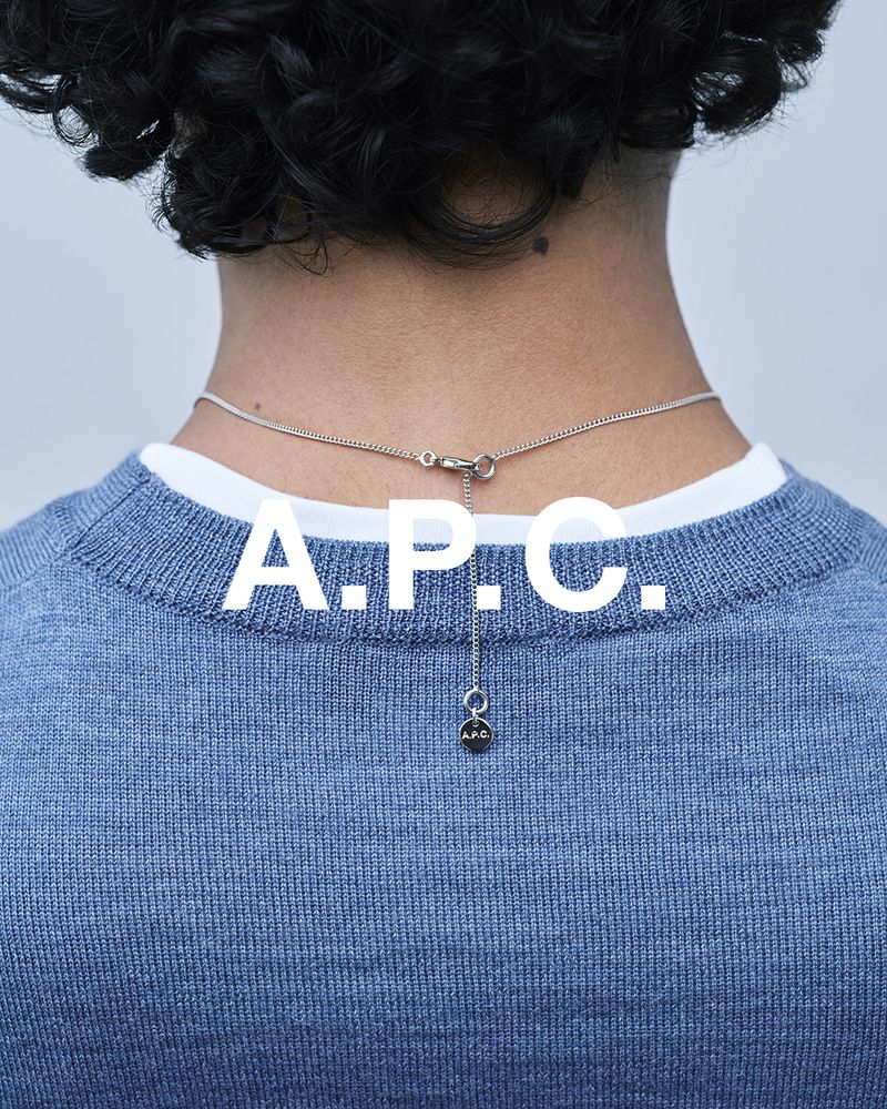 Back view of a person with curly hair wearing a blue sweater and silver necklace with the A.P.C. logo against a grey background.