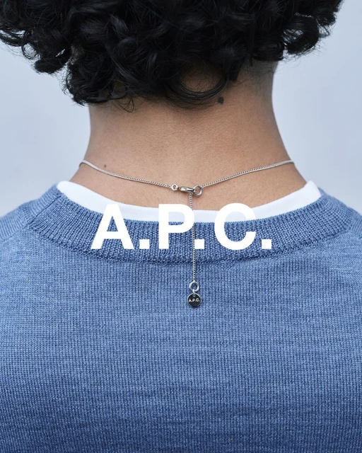 Back view of a person with curly hair wearing a blue sweater and silver necklace with the A.P.C. logo against a grey background.