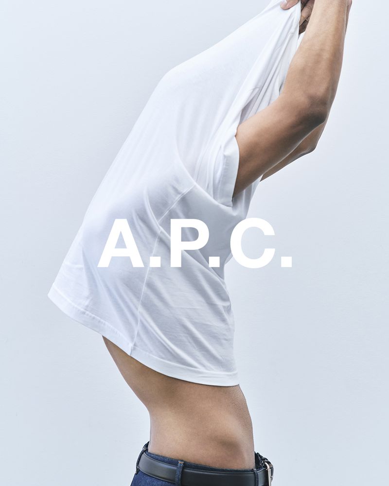 An adult male poses removing a white tee shirt against a neutral background in a studio fashion shot, emphasizing the clean aesthetic.