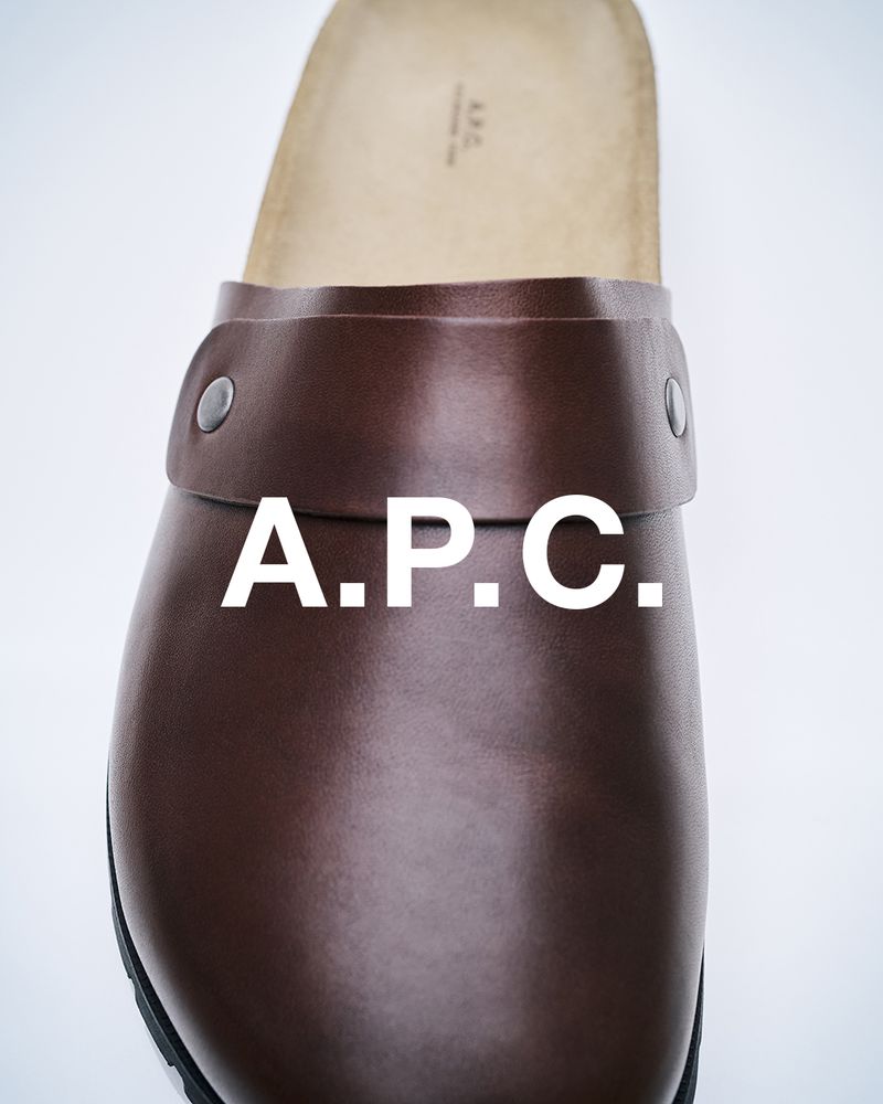 Close-up studio shot of a brown leather clog with a thick band and metal hardware, featuring the 'A.P.C.' brand logo in white.