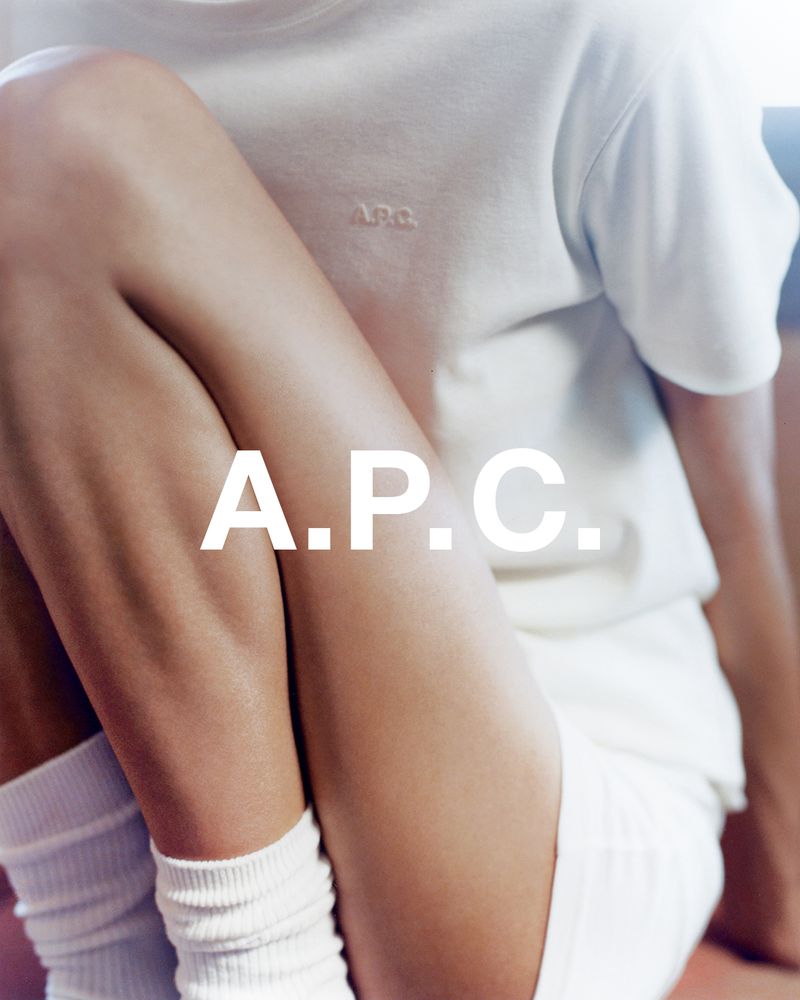 Close-up fashion shot of a woman wearing white T-shirt and shorts, sitting with legs crossed, featuring A.P.C. logo, and a soft, bright light.