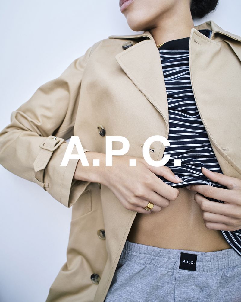 A person wearing a beige trench coat, striped shirt, and grey sweatpants poses against a white wall, showcasing fashion apparel.