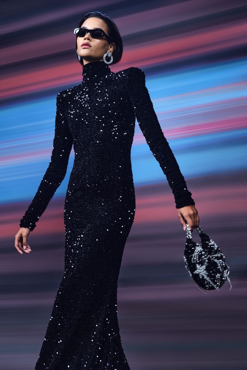 Model in black sequined turtleneck dress wearing sunglasses against a streaked colourful background holding an embellished handbag.