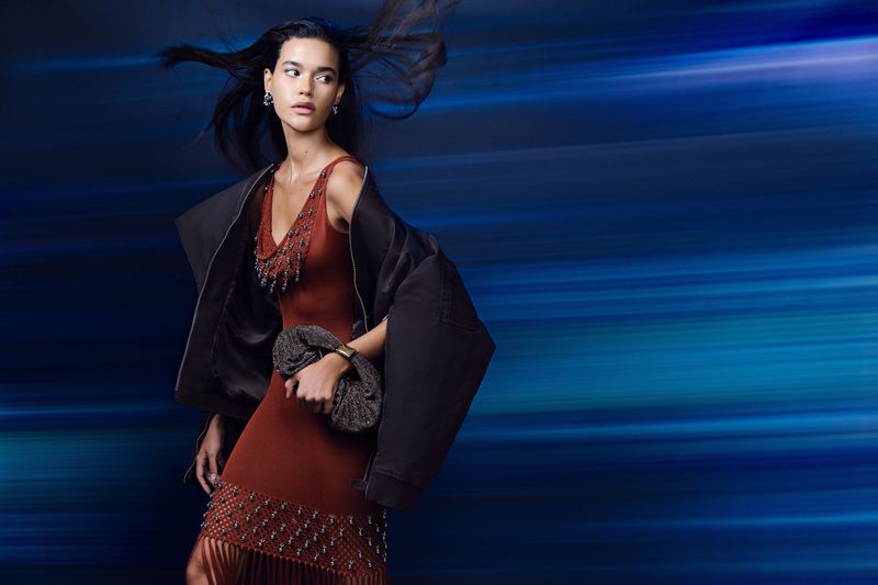 Fashion shot of a woman in a rust-colored dress and black jacket holding a clutch, set against a blurred blue background.