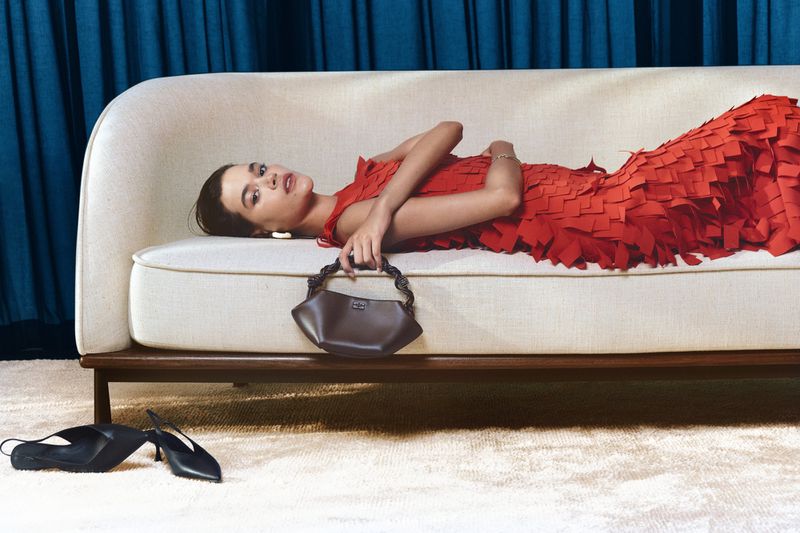 Model lying on a cream sofa wearing a textured red dress and holding a dark handbag for Harvey Nichols Dubai