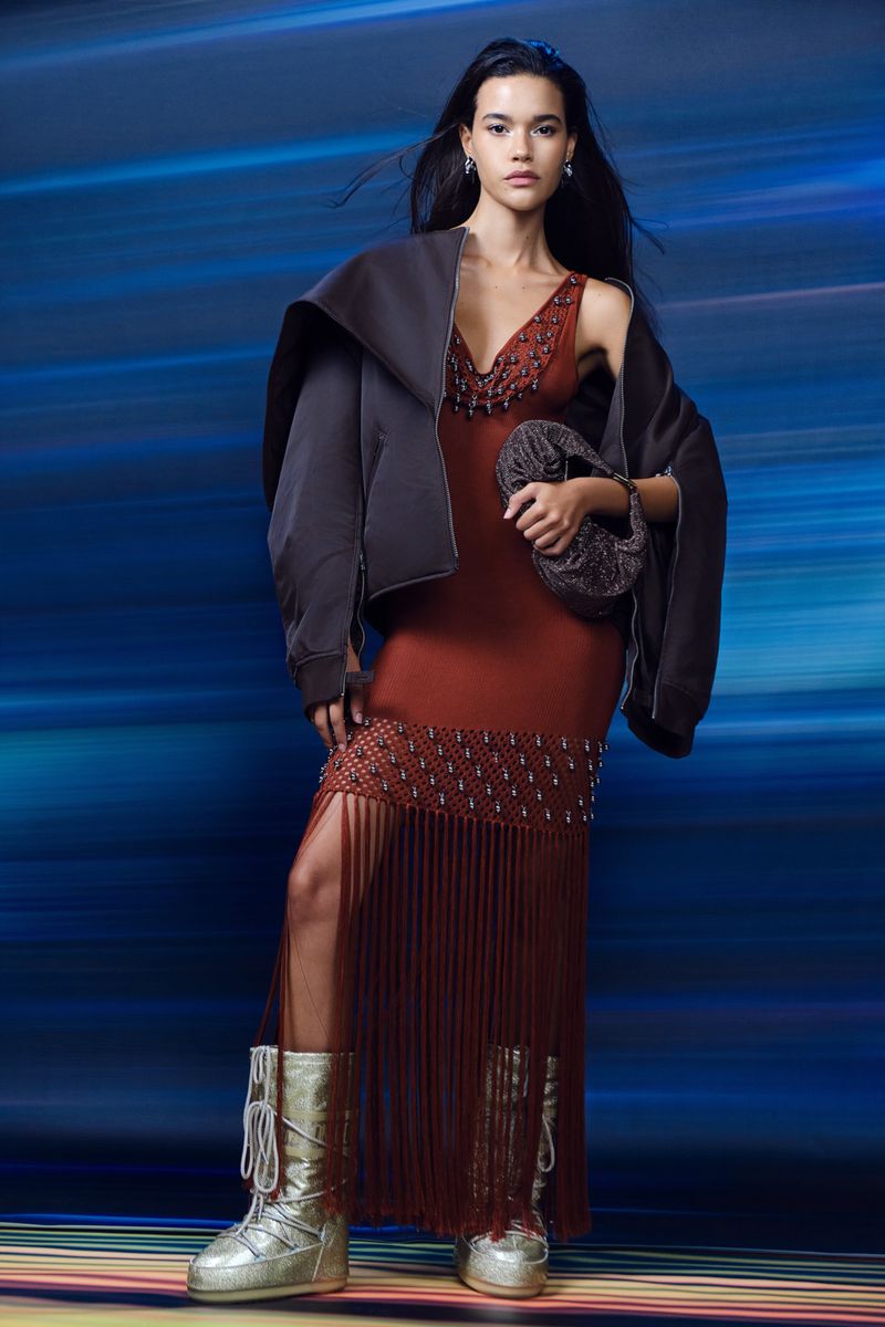 A woman models a brown beaded dress with fringe, paired with silver glittery boots and a dark jacket, against a blue gradient backdrop.