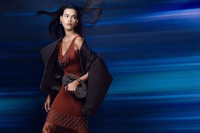 Model wearing a rust-colored dress and dark jacket holding a handbag against a blurred blue motion background